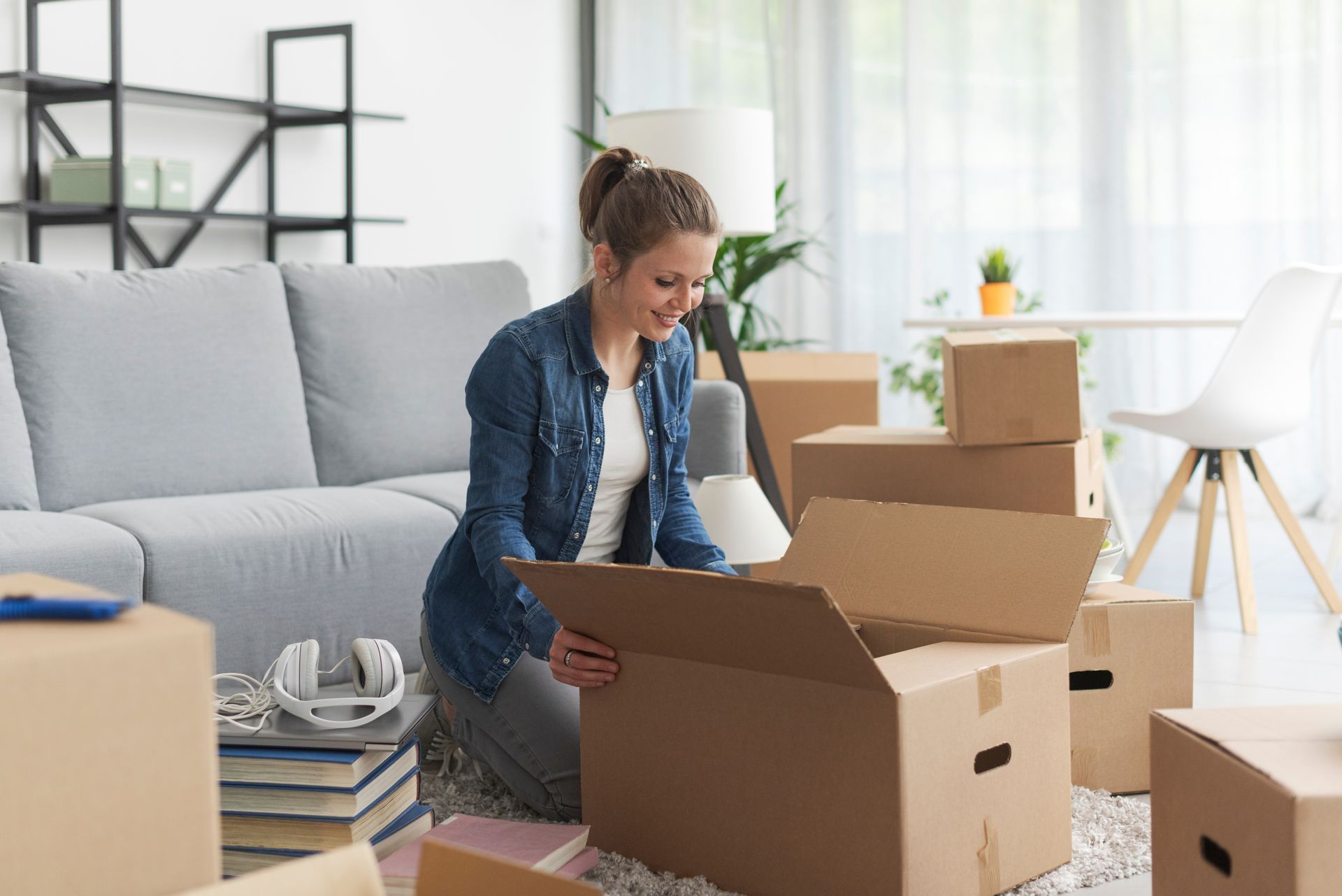 Woman kneeling, unpacking cardboard boxes in a light-filled living room; smiling.