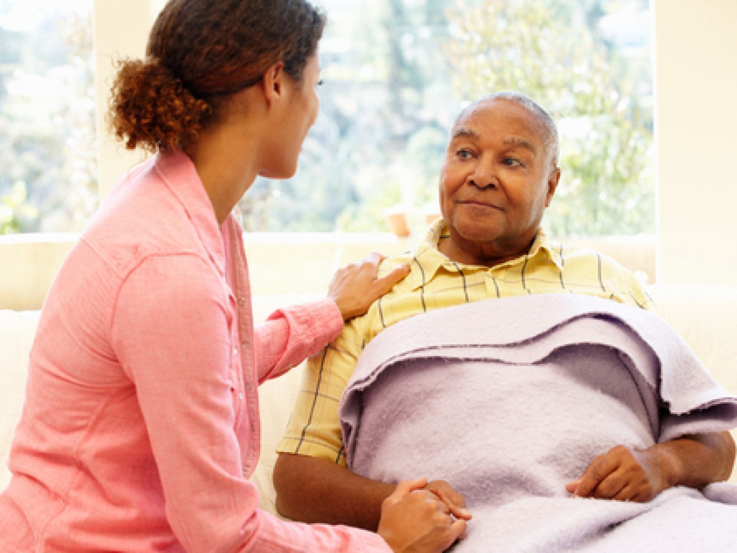 Woman comforts an elderly person, both seated. The woman wears pink, and the other person is under a blanket.