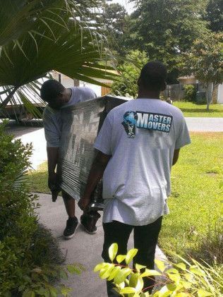 Two movers carrying a large appliance on a sidewalk in front of a house.