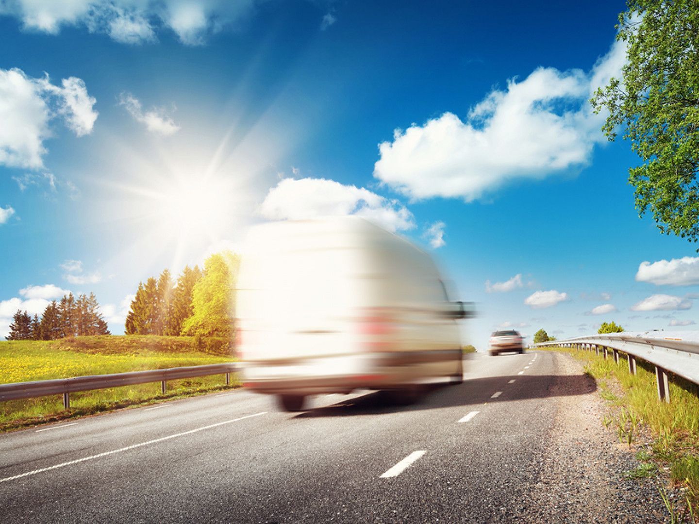 Blurred white delivery van on a highway under a bright sun, with blue sky and green landscape.