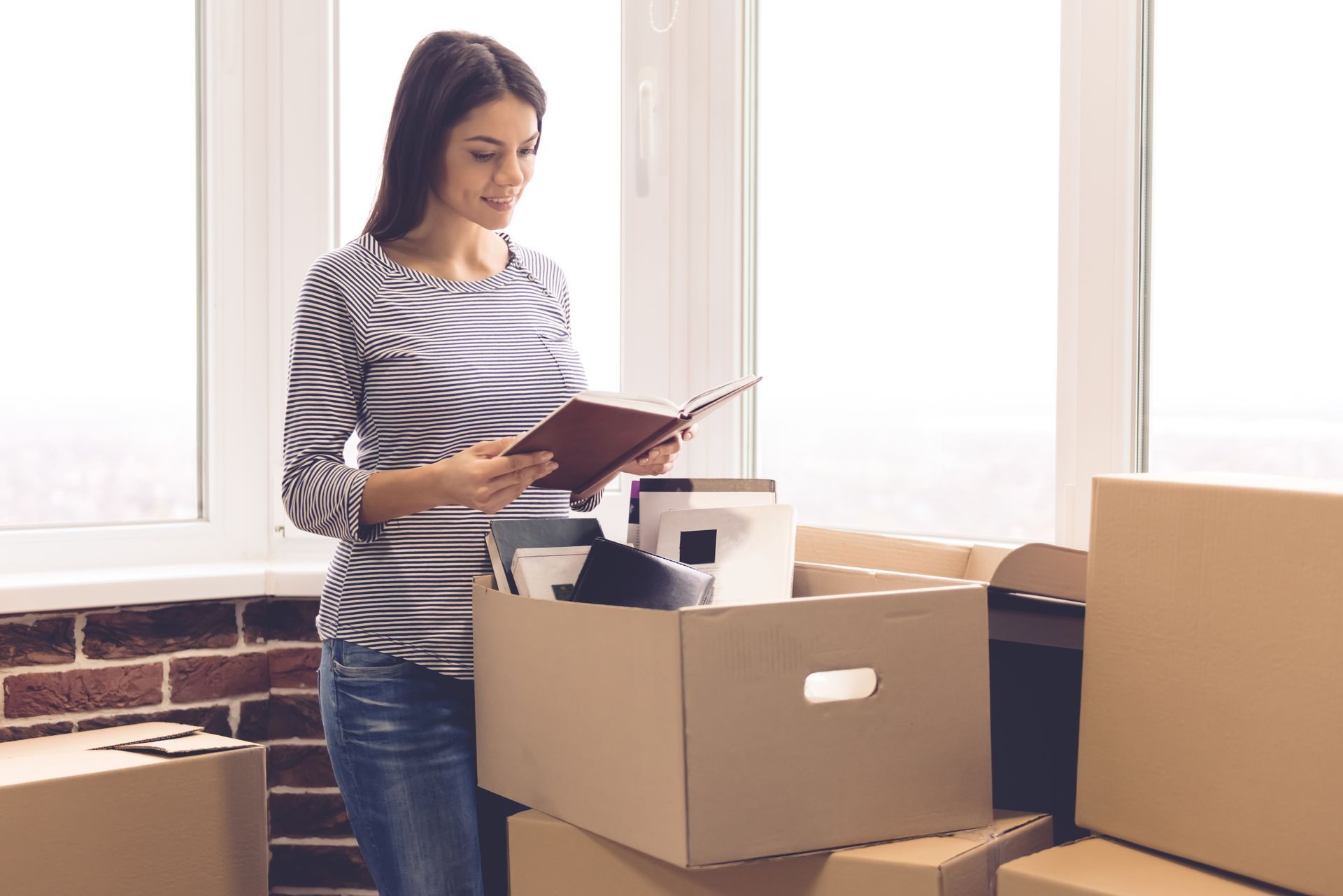 Woman looking at a book while unpacking boxes in a room with a window.