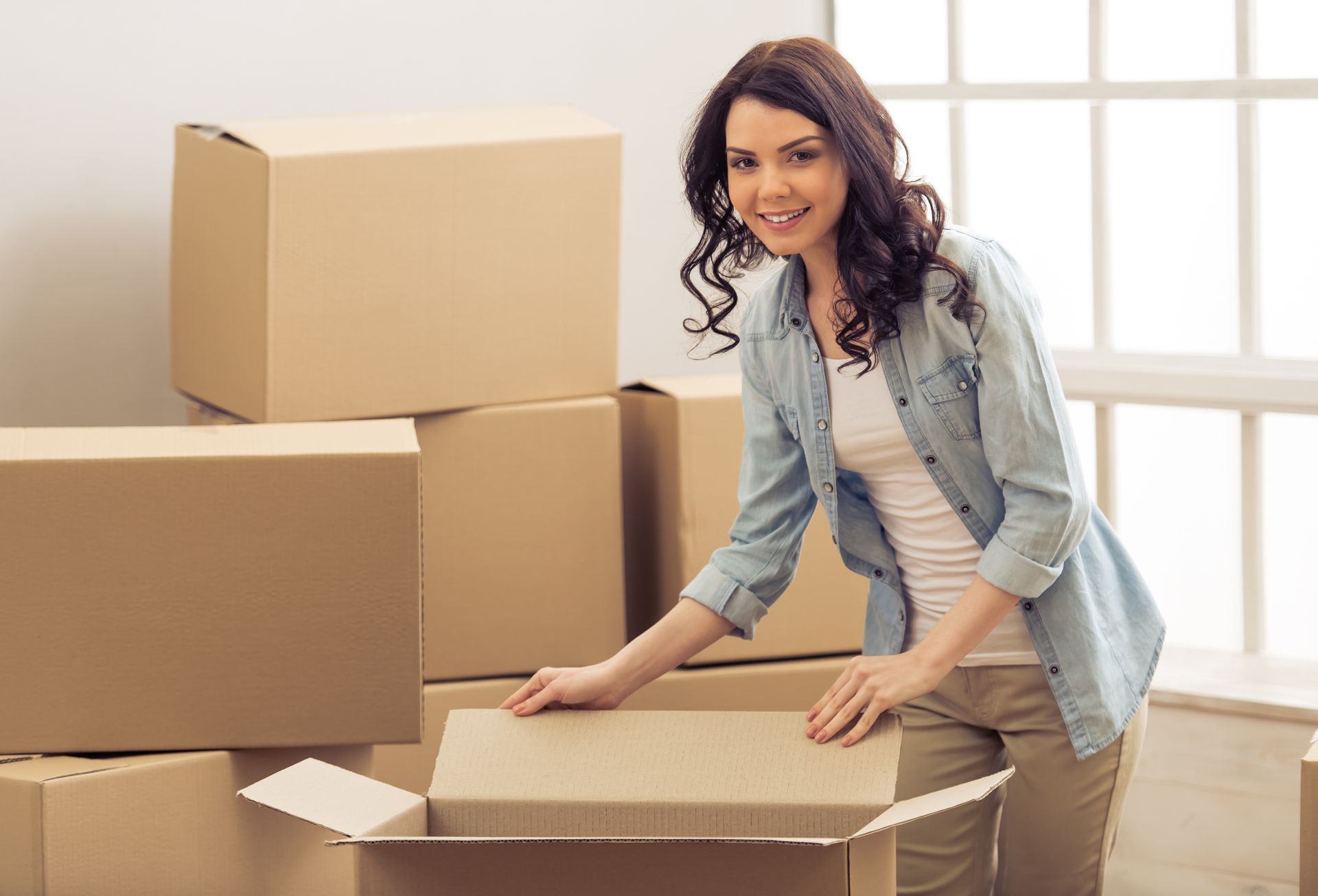 Woman smiling, opening a cardboard box. Surrounded by packed boxes in a room with a window.