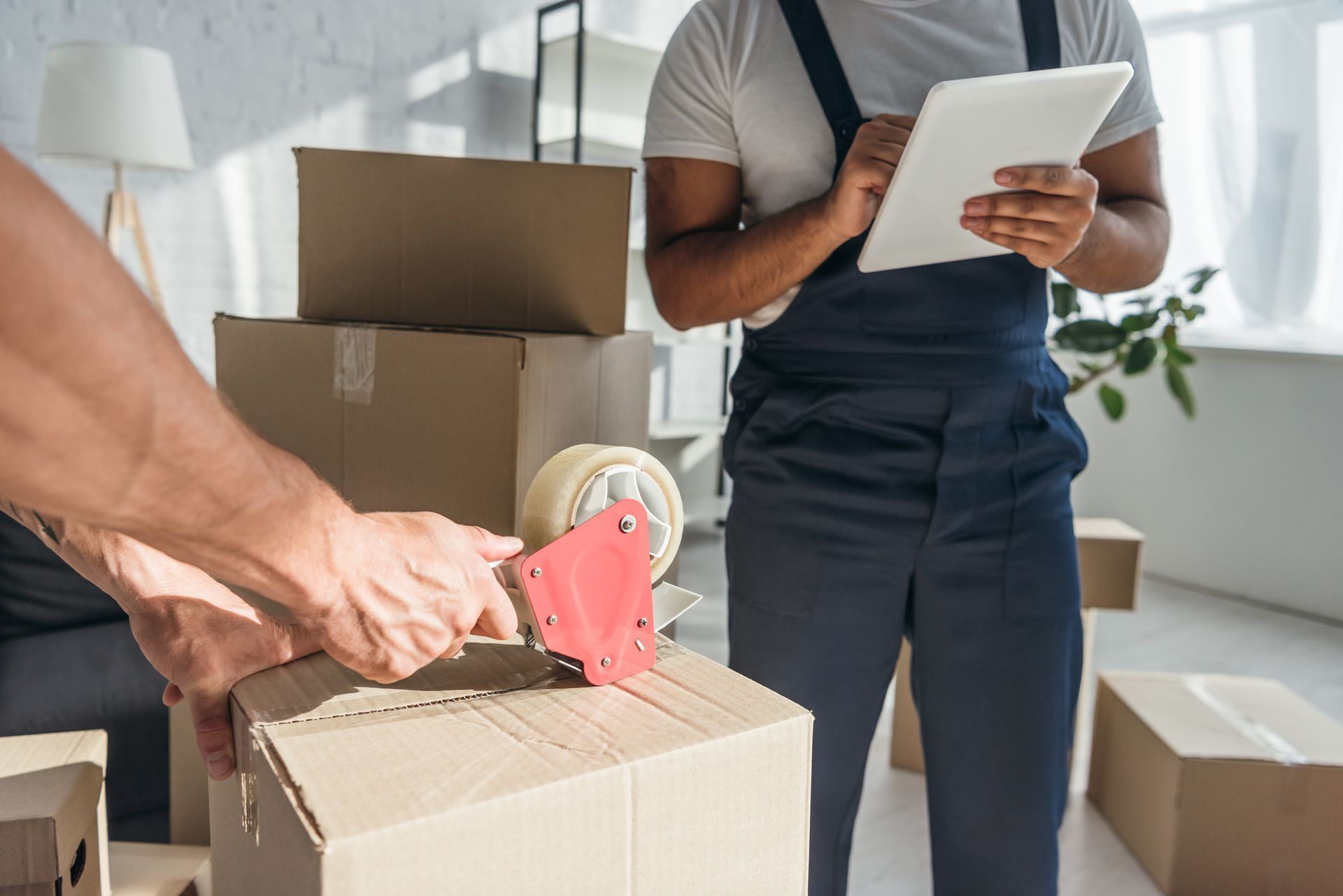 Movers packing boxes with tape, one using a tablet, in a room with sunlight.