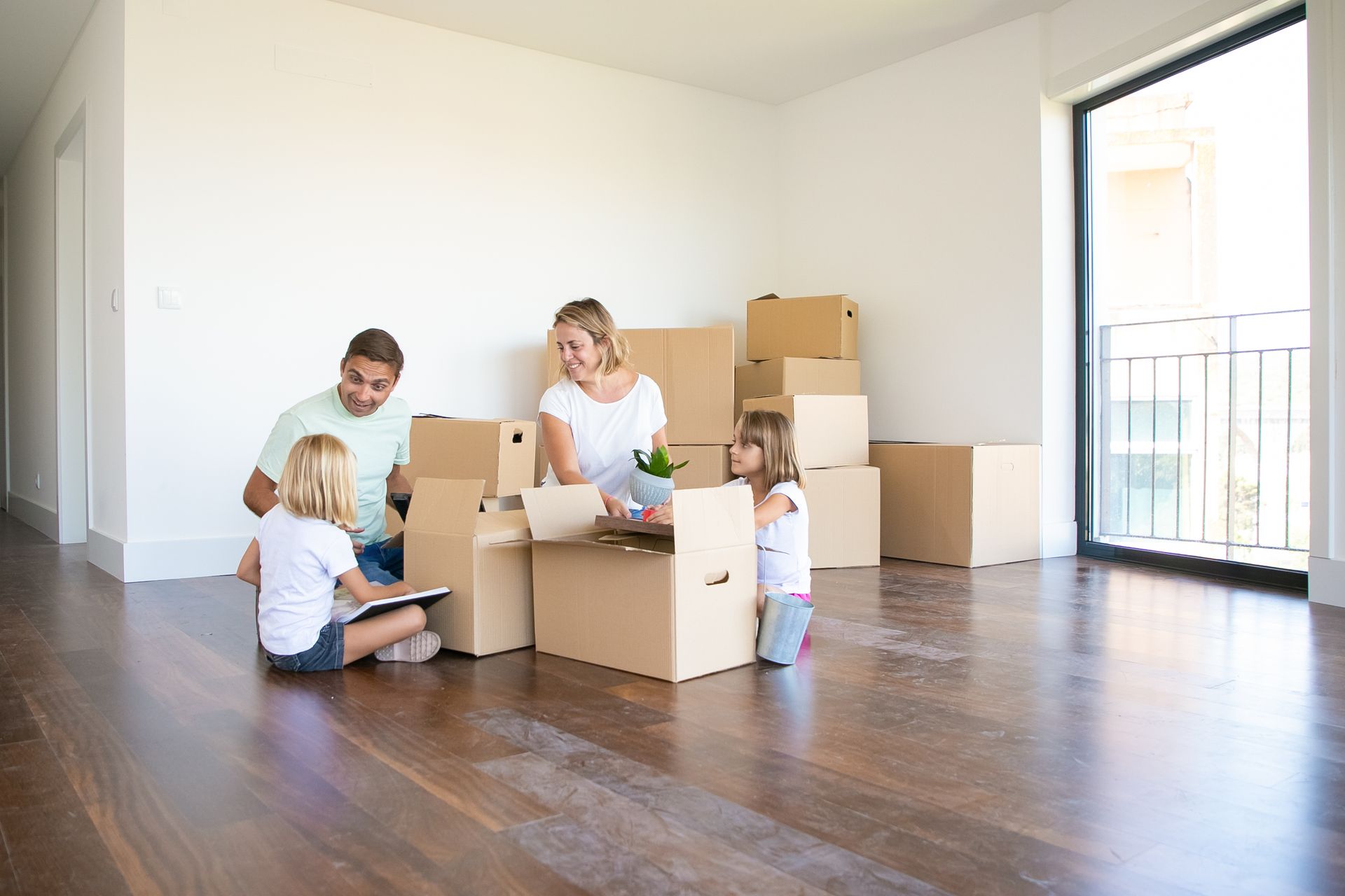 Family unpacking cardboard boxes in a new, empty room with a large window and hardwood floor.