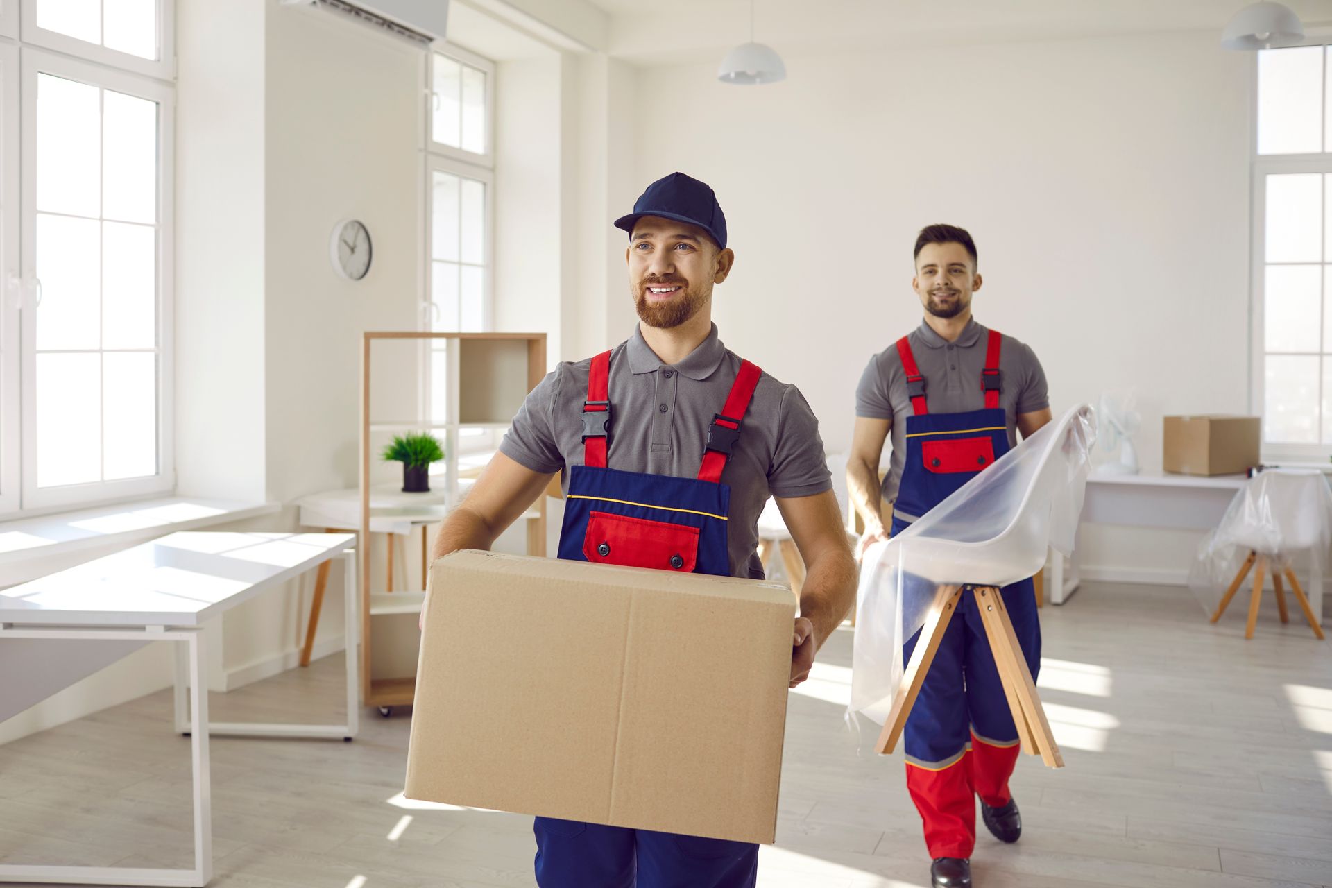 Two movers in red and blue uniforms carry a box and a chair in an empty room with sunlight.
