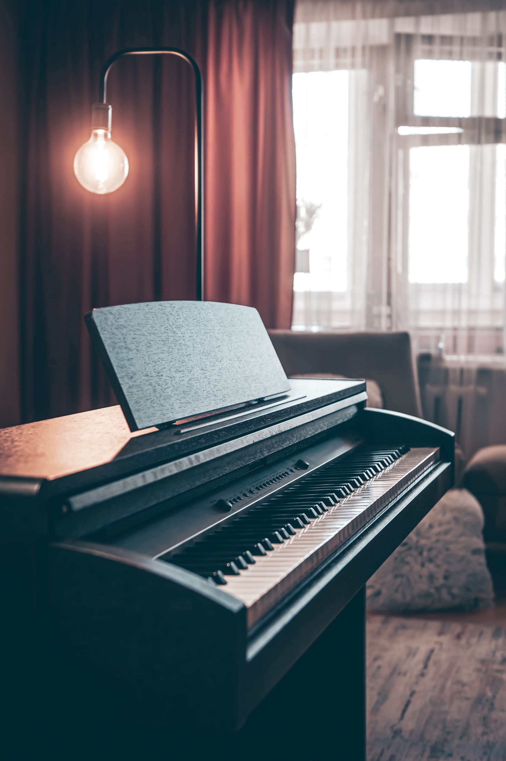 Piano with music stand and a lit bulb lamp in front of window with curtains.