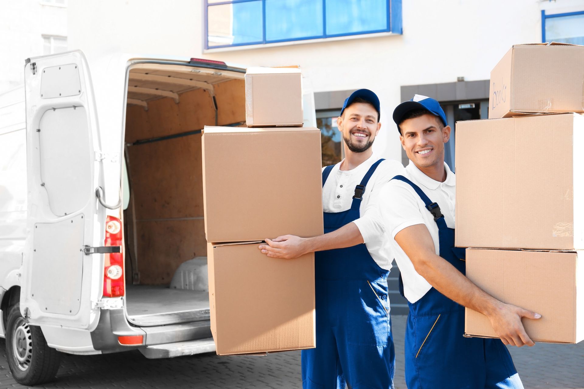 Two movers in blue overalls carrying cardboard boxes near a van, smiling.
